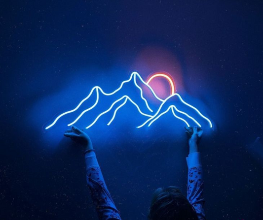 Person reaching toward a glowing neon wall art of mountain peaks and a setting sun, with a dark starry background.